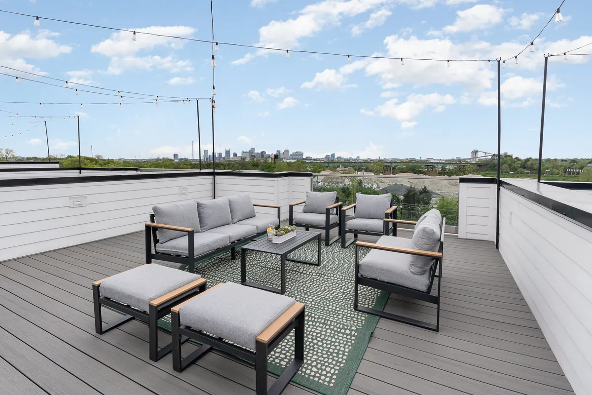 This photo shows a stylish rooftop outdoor seating area with comfortable lounge furniture and bistro string lights overhead. The city skyline is visible in the distance, enhancing the sense of an elevated urban retreat.