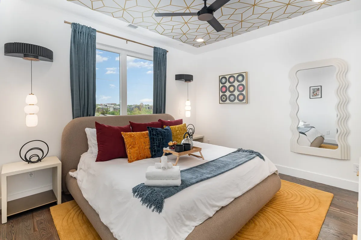 This image features a modern bedroom with a bed adorned with colorful pillows, a geometric ceiling design, a large window offering ample natural light, and a decorative mirror. The room exudes comfort and style, emphasizing privacy and boutique ambiance.