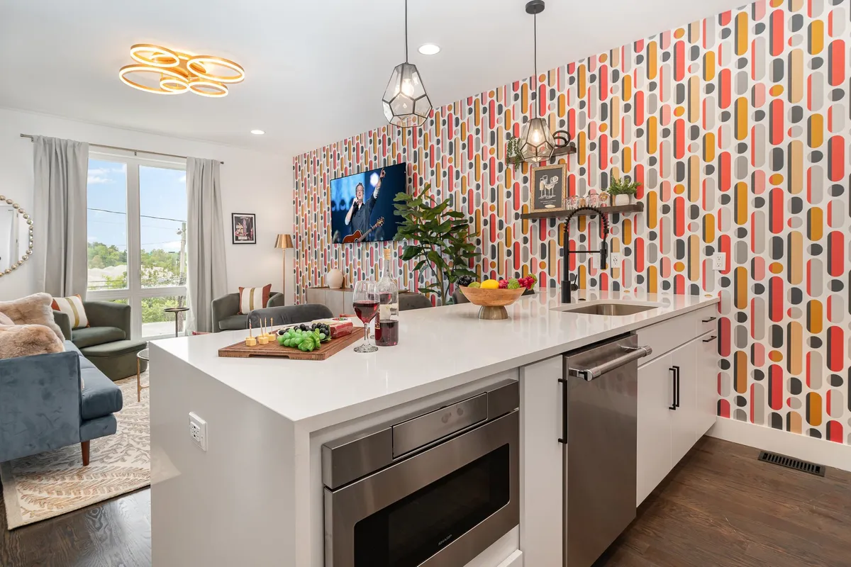 A bright, open-concept living, dining, and kitchen area featuring a colorful accent wall, a kitchen island with bar seating, a television displaying content, and lively greenery from a potted plant. The furniture and decor highlight a welcoming, cheerful atmosphere ideal for gatherings.