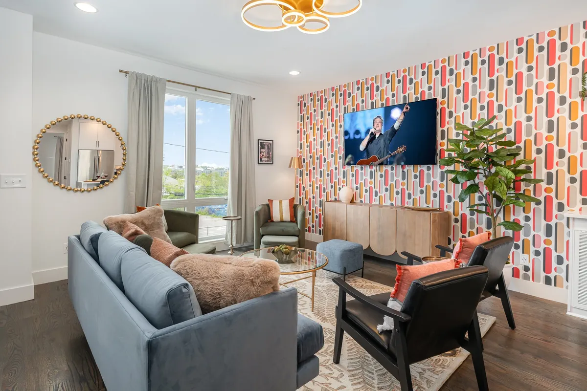 The image showcases a vibrant, modern living space with a colorful accent wall, plush stylish furniture, a large flat-screen TV, and a decorative mirror, all arranged to foster relaxation and social gatherings. The ambiance is lively yet cozy, ideal for groups to come together and unwind.