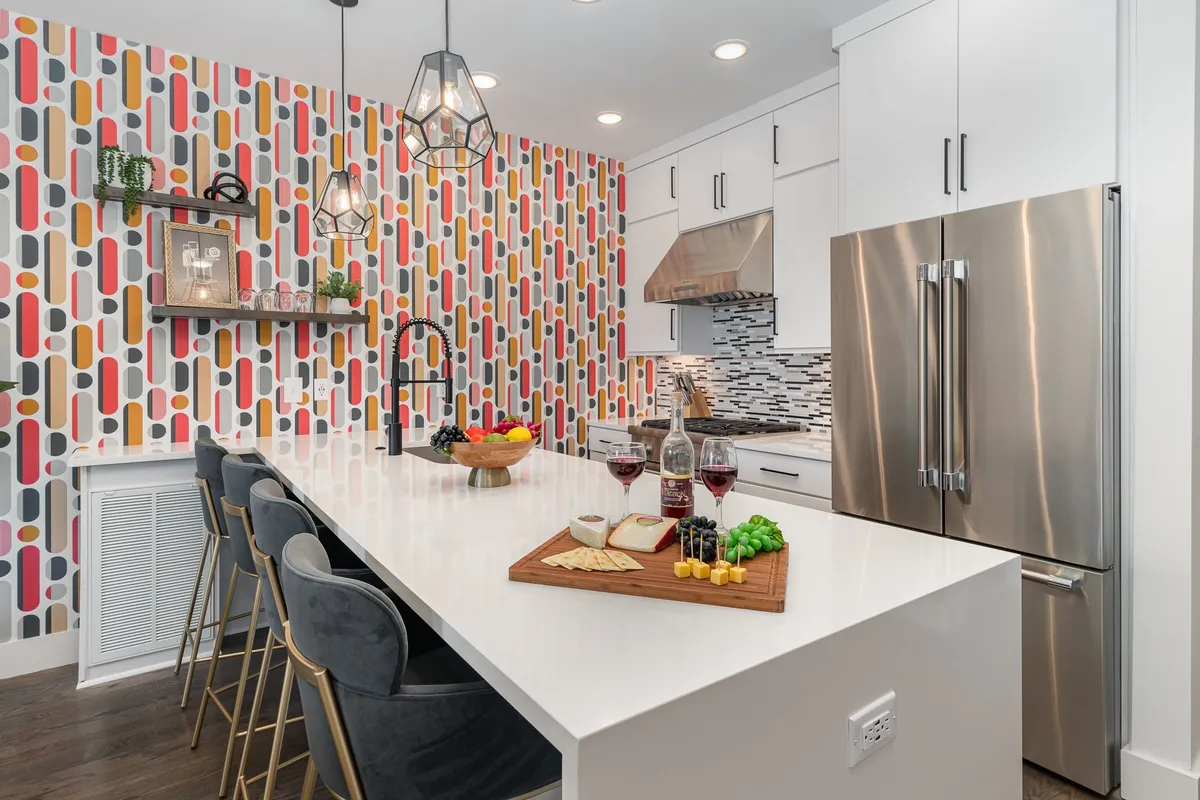 A cheerful and modern kitchen scene with colorful wallpaper, an island countertop set with food, stylish bar stools, and elegant hanging light fixtures. The setup encourages casual dining and lively conversation.