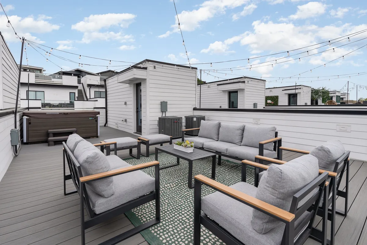A cozy rooftop corner featuring an inviting hot tub, a decorative outdoor rug, plush seating, and twinkling string lights. The setting is designed for relaxation and intimate conversations as the sun sets over the city.