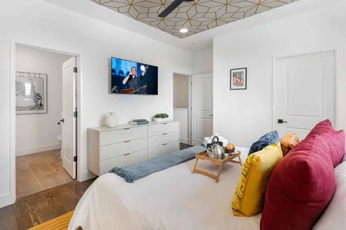 A king bedroom with a bed adorned with colorful pillows, a wall-mounted TV, artwork, and a nightstand with a tray for tea or coffee. The room is designed for comfort and relaxation, with a layout that emphasizes both style and function.